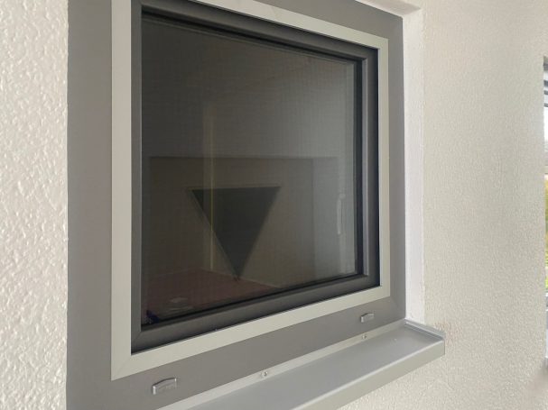 Ein quadratisches Fenster mit einem grauen Rahmen, das auf eine Wand blickt.