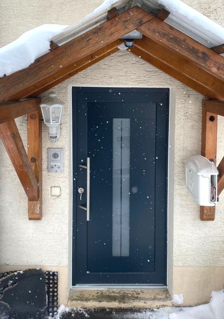 Moderne Eingangstür mit Glaselementen und Holzveranda, umgeben von Schnee.