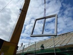 Ein Kran hebt einen quadratischen Rahmen über eine Baustelle mit Wolken im Hintergrund.