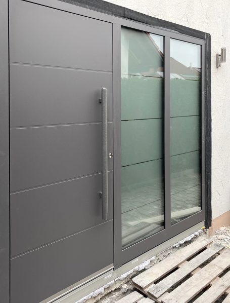Moderne graue Tür mit großen Glaselementen, daneben ein Holzuntergrund.