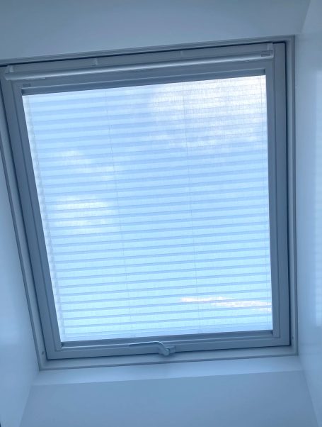 Blick auf einen hellen Himmel durch ein Dachfenster mit Jalousien.