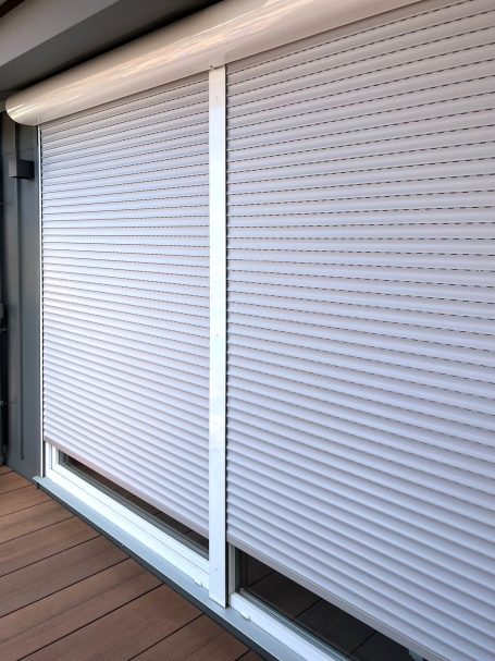 Weiße Rollläden, teilweise heruntergelassen, vor einem Fenster auf Holzboden.