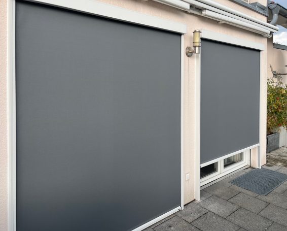 Graue Rollos an zwei Fenstern an einer hellen Wand.