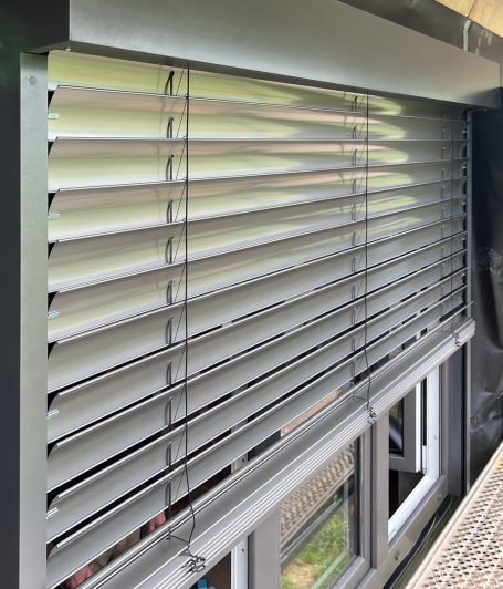 Blick auf eine graue Jalousie, die ein Fenster teilweise verdeckt.